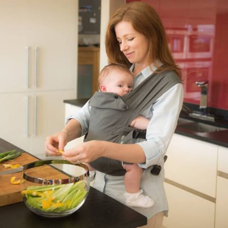 Woman prepares food while carrying baby in grey Izmi Baby Carrier