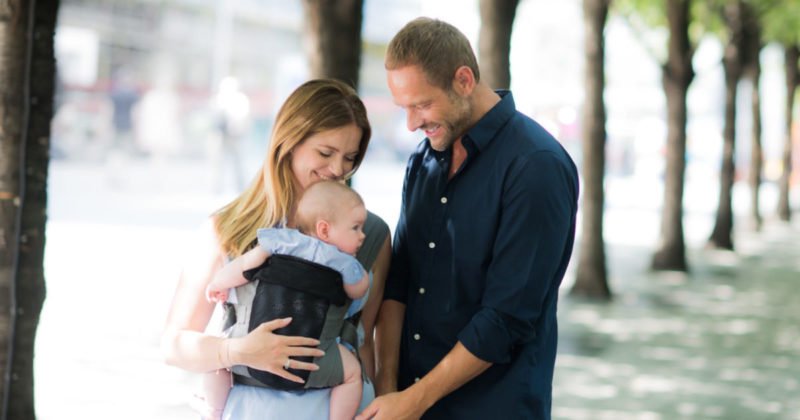 Woman carries baby in grey Izmi Breeze baby carrier while man smiles at baby