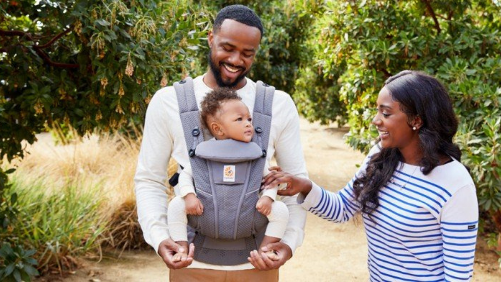 Man carries baby facing forwards in grey Ergobaby Omni Breeze carrier while woman smiles with them