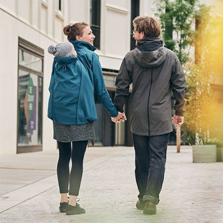 Woman carrying baby on her back while wearing the Mamalila Allrounder Softshell Babywearing Jacket in Teal. holding hands with man