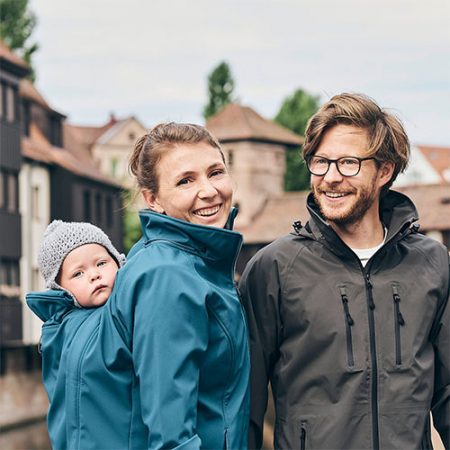 Woman carrying baby on her back while wearing the Mamalila Allrounder Softshell Babywearing Jacket in Teal