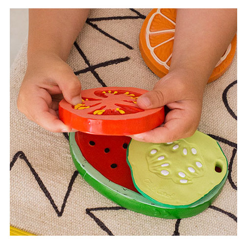 Baby hands hold tomato shaped rubber teething toy by Oli & Carol next to other fruit shaped toys