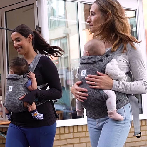 Two women walk while each carrying a baby facing towards them in the Carifit+ baby carrier